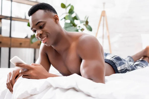 Homem americano africano alegre usando smartphone na cama em primeiro plano desfocado — Fotografia de Stock