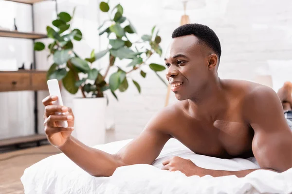 Sonriendo afican americano hombre tomando selfie en teléfono inteligente en la cama durante la mañana - foto de stock