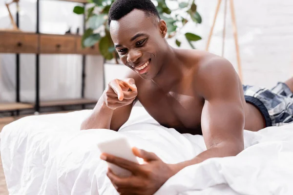 Fröhlicher afrikanisch-amerikanischer Mann zeigt auf Smartphone im verschwommenen Vordergrund auf Bett — Stockfoto