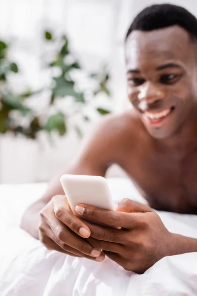 Smartphone in hands of smiling african american man on white bedding on blurred background — Stock Photo