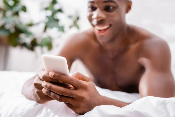 Смартфон в руках веселого афроамериканського чоловіка на розмитому фоні в спальні. — Stock Photo