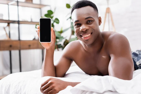 Hombre afroamericano sin camisa mostrando teléfono inteligente con pantalla en blanco en la cama - foto de stock