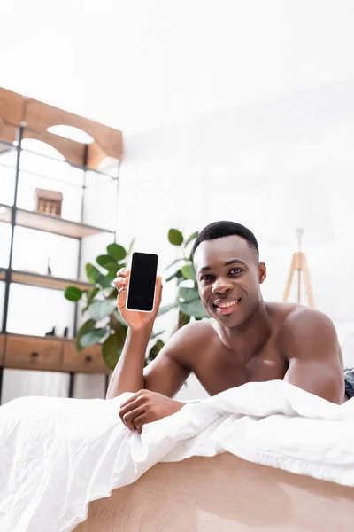 Afrikanischer Mann lächelt und zeigt Smartphone mit leerem Bildschirm auf dem Bett — Stockfoto