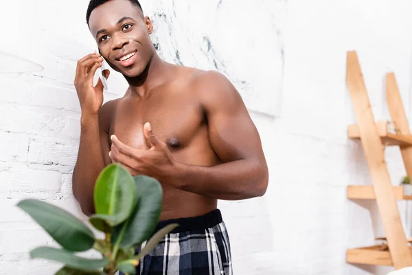 Африканский мужчина без рубашки разговаривает на смартфоне рядом с заводом на размытом переднем плане дома — стоковое фото