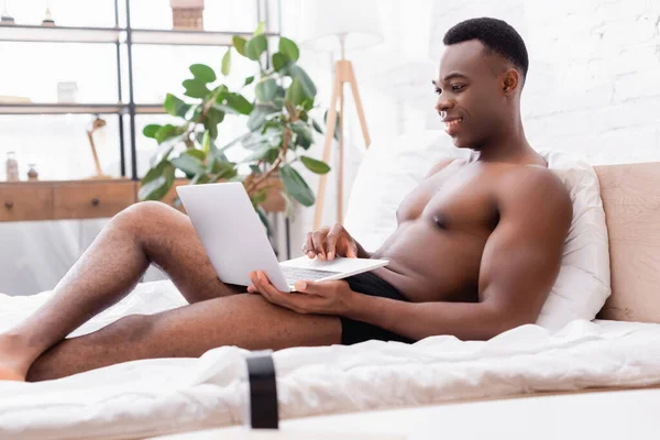 Усміхнений афроамериканський чоловік в трусиках, використовуючи ноутбук на ліжку на розмитому передньому плані — стокове фото