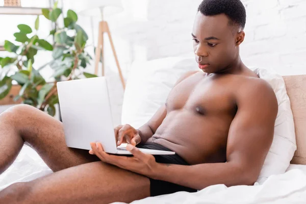 Hombre afroamericano sin camisa usando el ordenador portátil mientras está acostado en la cama - foto de stock