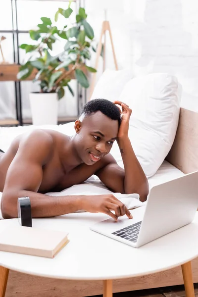 Shirtless african american man using laptop near clock and book on coffee table in bedroom — Stock Photo