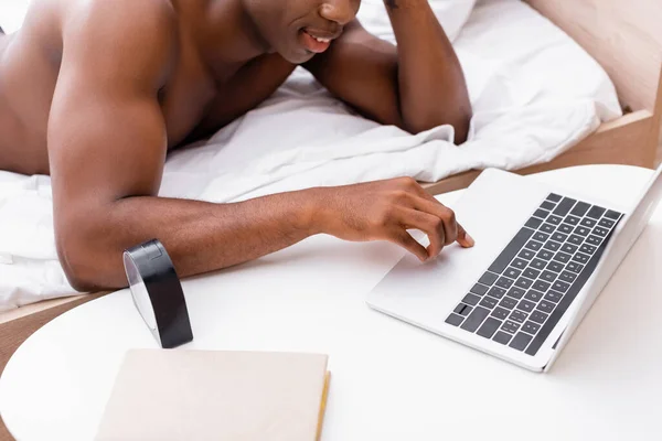 Vista recortada del reloj y libro en la mesa cerca del hombre afroamericano sonriente utilizando el ordenador portátil en la cama sobre fondo borroso - foto de stock