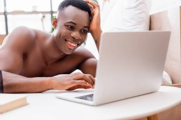 Joyeux homme afro-américain utilisant un ordinateur portable au premier plan flou dans la chambre — Photo de stock