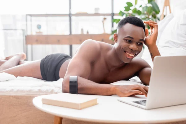 African american man in underpants using laptop while lying on bed near book and clock on blurred foreground — Stock Photo