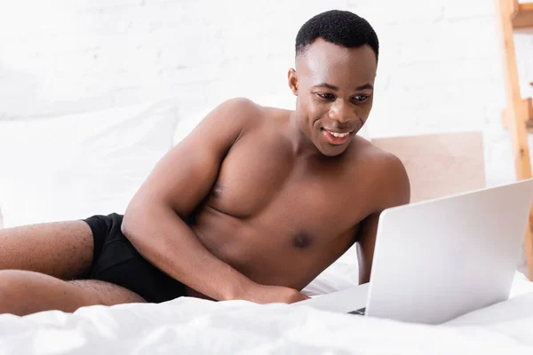 Улыбающийся африканский американец в нижнем белье с помощью ноутбука на размытом переднем плане на кровати — стоковое фото
