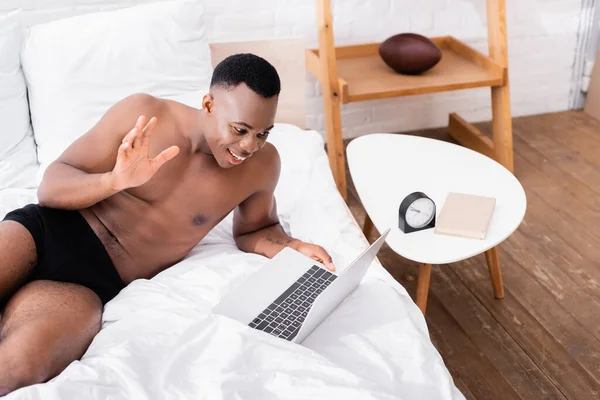 Cheerful shirtless african american man waving hand during video call on laptop near book and clock on coffee table — Stock Photo