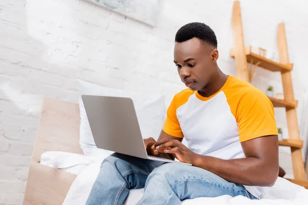 Молодой африканский американец, работающий на ноутбуке, сидя дома на кровати — стоковое фото