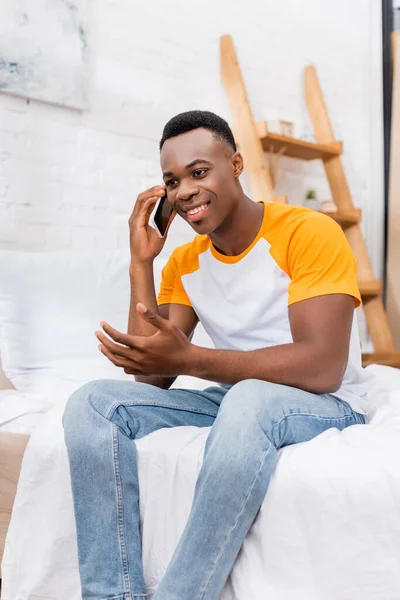 Positivo afroamericano hombre hablando en el teléfono inteligente mientras está sentado en la cama por la mañana - foto de stock