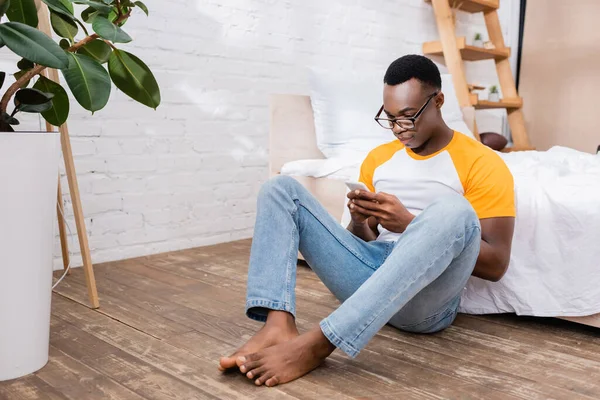 Barefoot african american man in eyeglasses using smartphone on floor in bedroom — Stock Photo