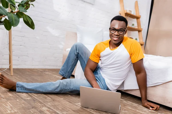 Lächelnder afrikanisch-amerikanischer Mann schaut in Kamera, während er Laptop auf dem Fußboden im Schlafzimmer benutzt — Stockfoto