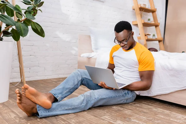 Barefoot african american freelancer using laptop while sitting near bed and plant at home — Stock Photo