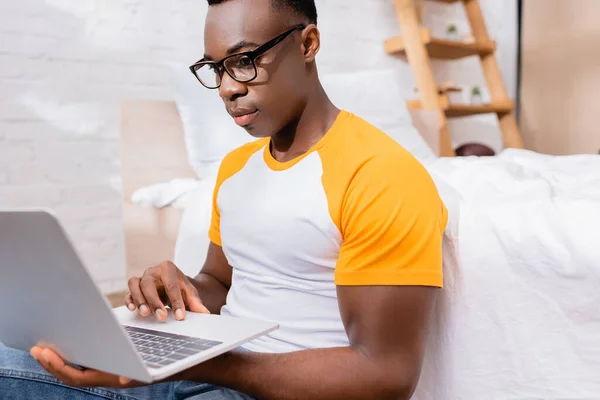 African american freelancer in eyeglasses using laptop on blurred foreground in bedroom — Stock Photo