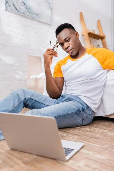 Pensive african american man holding eyeglasses near laptop on blurred foreground on floor — Stock Photo