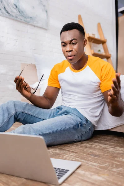 Angry african american freelancer holding eyeglasses near laptop on blurred foreground on floor in bedroom — Stock Photo