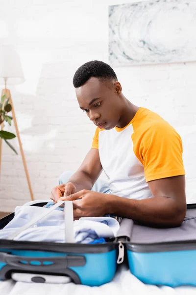 Young african american man holding belts of suitcase with clothes on bed on blurred foreground — Stock Photo