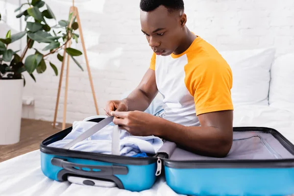 Hombre afroamericano recogiendo maleta mientras está sentado en la cama en casa - foto de stock