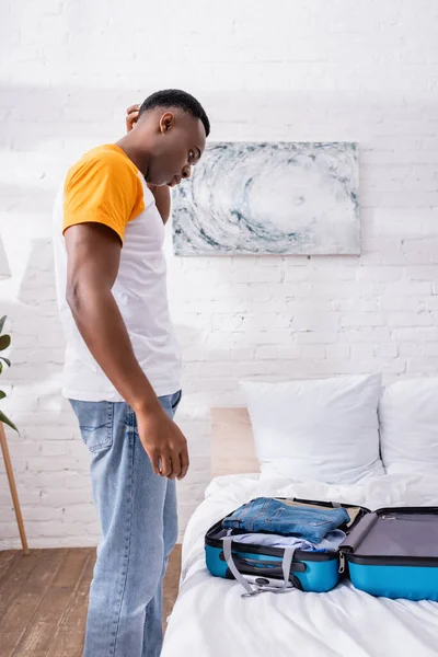 Vista lateral del hombre afroamericano reflexivo mirando la maleta con ropa en la cama — Stock Photo
