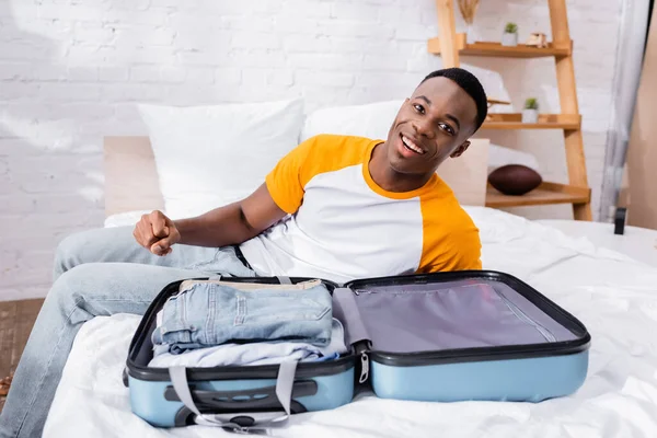 Joyeux homme afro-américain regardant la caméra près de la valise avec des vêtements sur le lit à la maison — Photo de stock