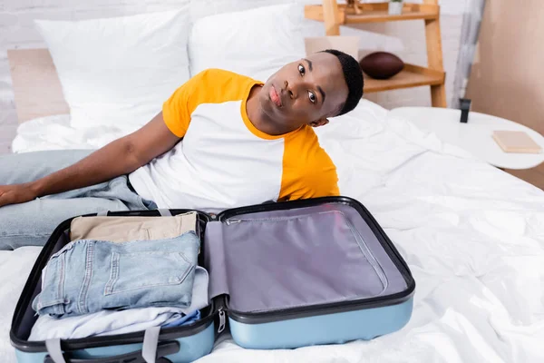 Valise avec des vêtements près de l'homme afro-américain regardant la caméra sur le lit — Photo de stock
