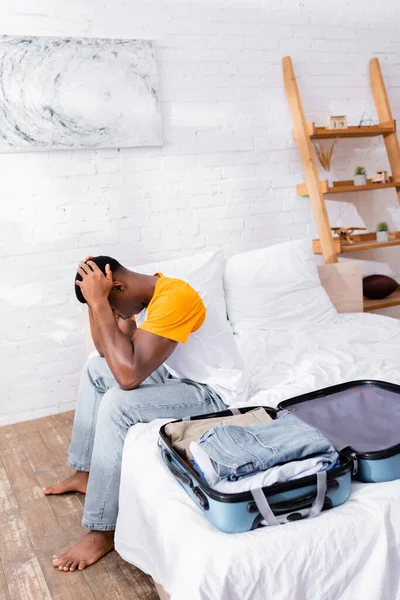 Dissatisfied african american man sitting near suitcase with clothes on bed — Stock Photo