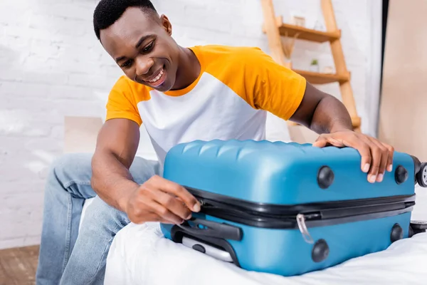 Homme afro-américain positif verrouillant sa valise au premier plan flou sur le lit — Photo de stock