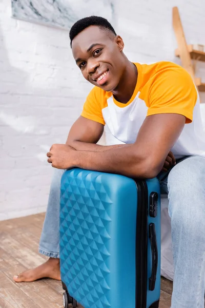 Joyeux homme afro-américain regardant la caméra près de la valise à la maison — Photo de stock