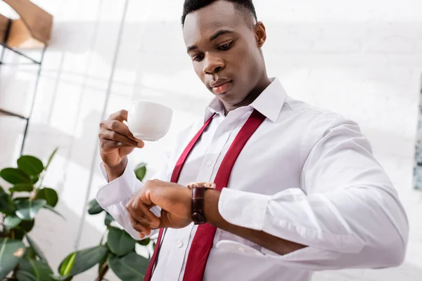 African american businessman holding coffee cup and looking at wristwatch on blurred foreground at home — Stock Photo