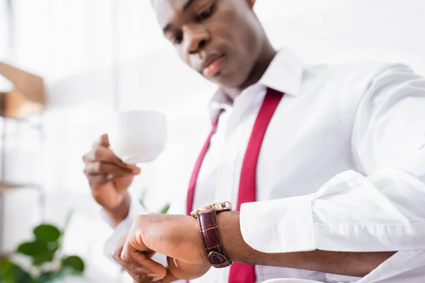 Reloj de pulsera en brazo de hombre de negocios afroamericano con taza de café sobre fondo borroso en casa - foto de stock