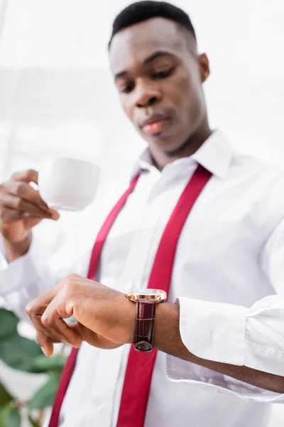 Wristwatch on hand of african american man in shirt and tie holding cup of coffee on blurred background at home — Stock Photo