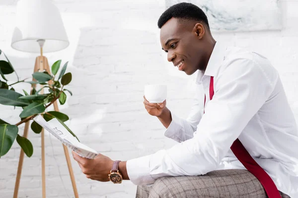 Homme d'affaires afro-américain souriant avec tasse de café lecture journal à la maison — Photo de stock