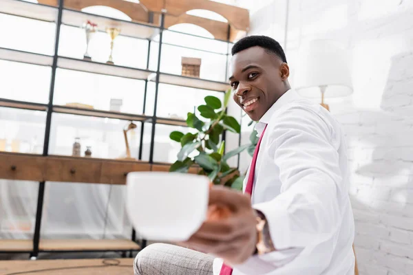 Cheerful african american businessman holding cup on blurred foreground at home — Stock Photo