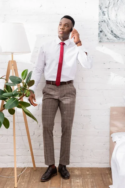 Africano americano empresário falando no smartphone perto da cama e planta em casa — Fotografia de Stock