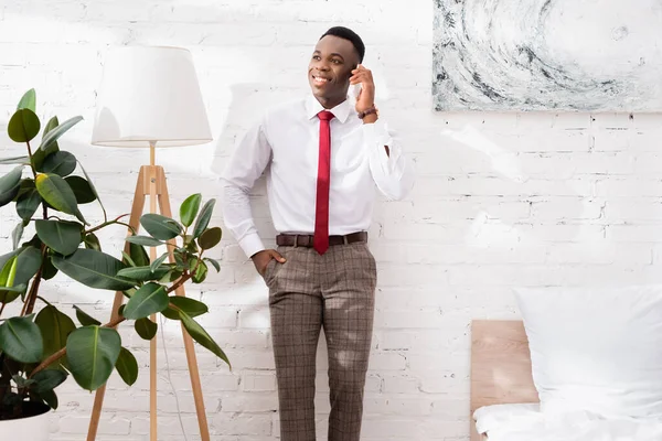 Smiling african american businessman talking on smartphone near floor lamp in bedroom — Stock Photo