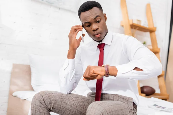 Afrikanischer Geschäftsmann spricht auf Smartphone und schaut auf Armbanduhr, während er auf verschwommenem Hintergrund im Bett sitzt — Stockfoto