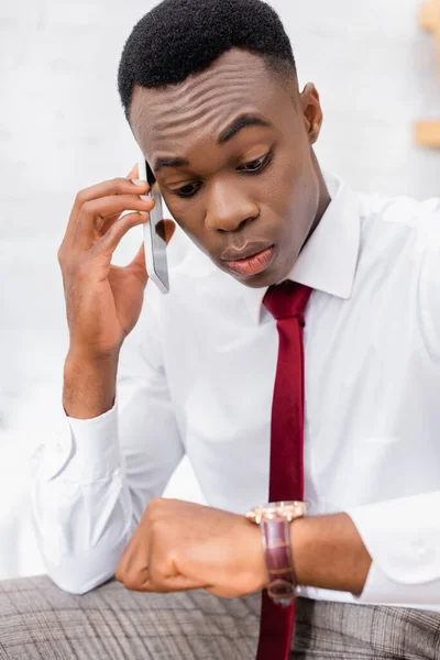 Hombre de negocios afroamericano en camisa hablando en smartphone y mirando el reloj de pulsera en primer plano borroso - foto de stock
