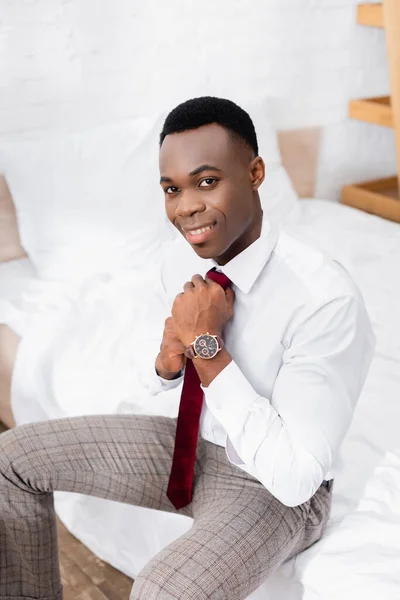 Alegre hombre de negocios afroamericano con corbata roja mientras está sentado en la cama en casa - foto de stock