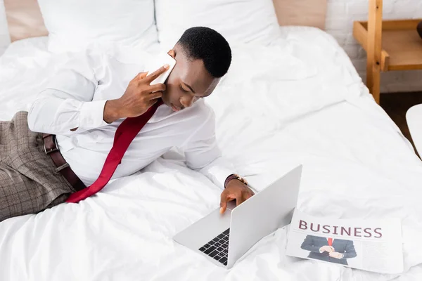 African american businessman talking on smartphone and using laptop near newspaper on bed — Stock Photo