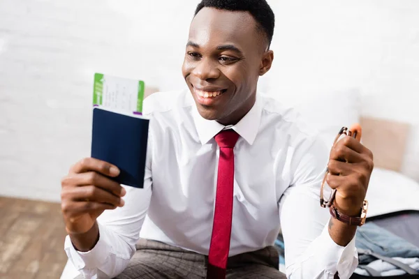 Uomo d'affari afroamericano sorridente con occhiali e passaporto con biglietto aereo in primo piano sfocato — Foto stock