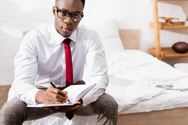 Empresário afro-americano em óculos olhando para a câmera enquanto escrevia no caderno em primeiro plano desfocado na cama — Fotografia de Stock