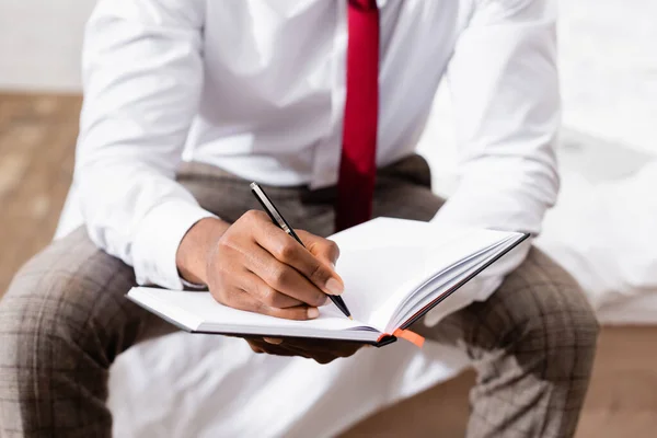 Cropped view of african american businessman writing on notebook on blurred background on bed — Stock Photo