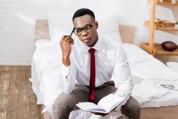 Offensiv afrikanisch-amerikanischer Geschäftsmann in Brille mit Stift und Notizbuch auf dem Bett vor verschwommenem Hintergrund — Stockfoto