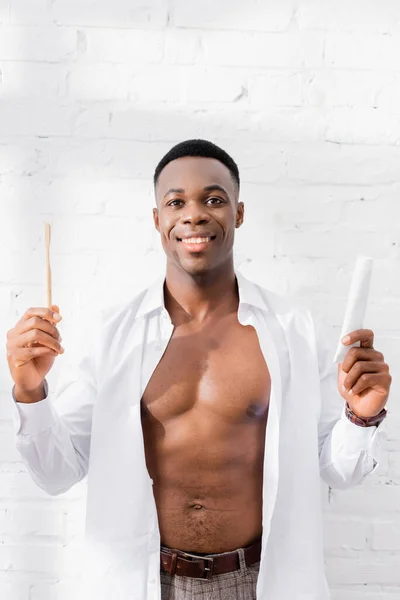 Muscular african american man in shirt smiling while holding toothpaste and toothbrush at home — Stock Photo