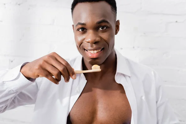 Smiling african american man in shirt holding toothbrush with toothpaste at home — Stock Photo