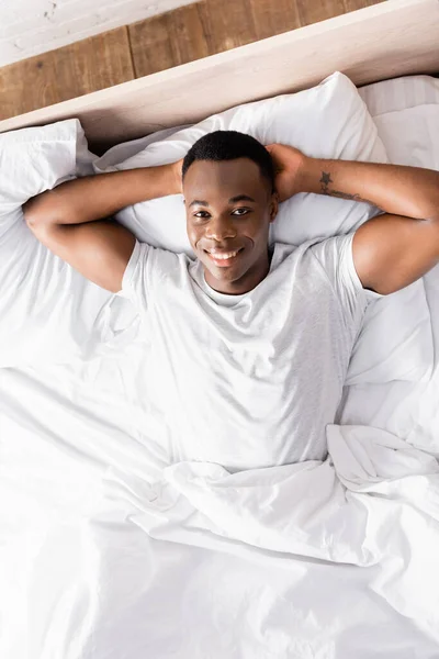 Blick von oben auf tätowierten Afroamerikaner, der auf dem Bett in die Kamera lächelt — Stockfoto